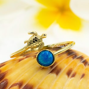 May include: Gold ring with a blue opal stone and a small turtle design. The ring is on a seashell background.