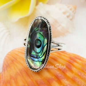 May include: Oval shaped abalone shell ring with a silver band. The ring is set on a seashell with a white background.