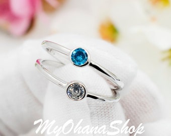 May include: Two silver rings with small gemstones. The top ring has a light blue gemstone and the bottom ring has a clear gemstone. The rings are on a white surface.