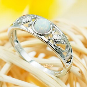 May include: A silver ring with a white opal gemstone and a design of three sea turtles. The ring is on a woven background.