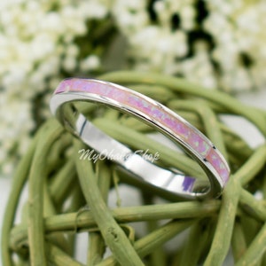 May include: A thin silver band ring with a pink opal inlay. The ring is sitting on a woven green base.