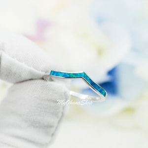 May include: A silver ring with a chevron-shaped band inlaid with vibrant blue-green opal stones. The ring is held against a soft, blurred background, highlighting the jewelry's design and color. The ring is a delicate piece of jewelry.