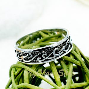 Könnte beinhalten: Ein silberner Ring mit einem Wellendesign. Der Ring liegt auf einem grünen Hintergrund.