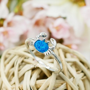 May include: A silver crab ring with a blue opal stone. The ring features a crab design with detailed claws and a textured band. The ring is set against a blurred background of pink flowers and a woven surface. The text "MyOhana Shop" is visible.