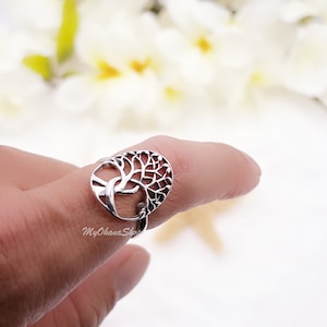 May include: A silver ring with a tree of life design. The ring's oval face displays the tree's branches and roots in detail. The ring is worn on a finger, with a blurred floral background. The ring is a piece of jewellery.