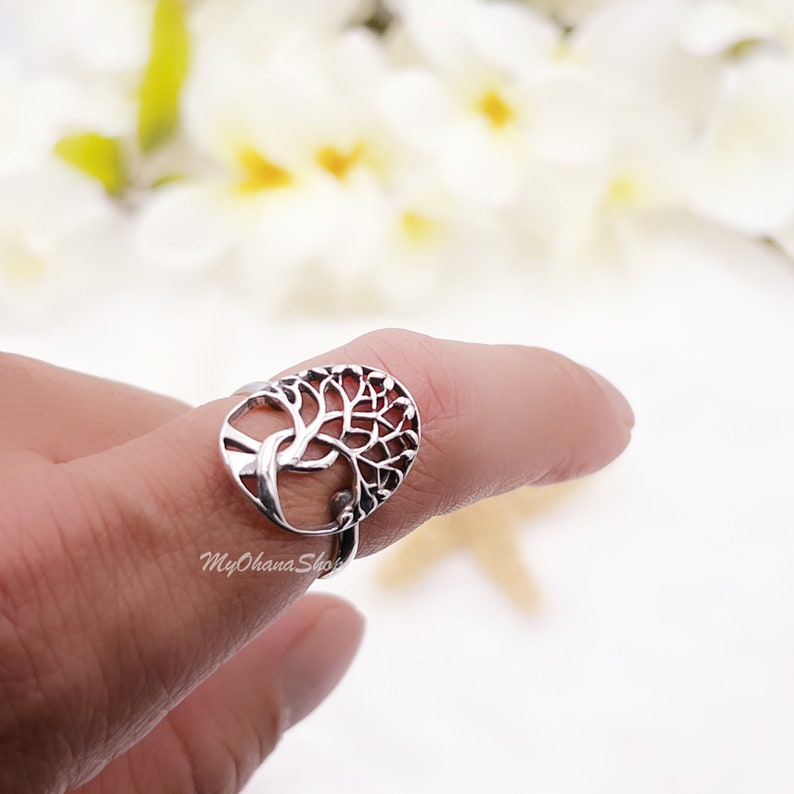 May include: A silver ring with a tree of life design. The ring's oval face displays the tree's branches and roots in detail. The ring is worn on a finger, with a blurred floral background. The ring is a piece of jewellery.
