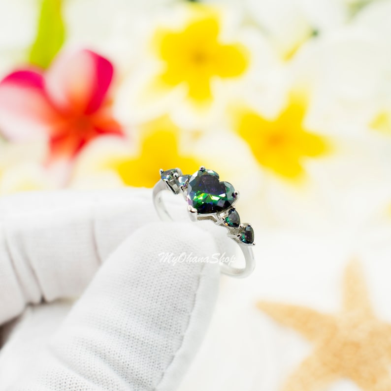 Op de afbeelding: Een zilveren ring met een grote hartvormige groene en zwarte edelsteen, omringd door kleinere edelstenen. De ring ligt op een wit oppervlak.