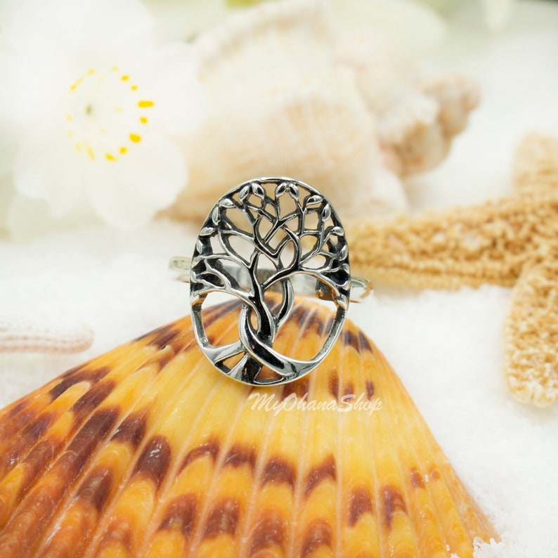 May include: A silver ring with an oval tree of life design. The ring is set against a seashell and beach-themed elements. The ring's design is intricate and detailed. The ring is likely made of sterling silver.