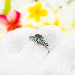 Op de afbeelding: Een zilveren ring met een grote hartvormige groene edelsteen, omringd door kleinere hartvormige edelstenen.