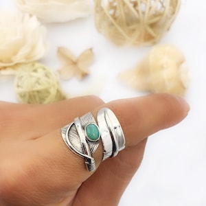 Op de afbeelding: Twee zilveren ringen aan een vinger. De bovenste ring heeft een zilveren verenontwerp met een turquoise steen. De onderste ring is een eenvoudige zilveren band.
