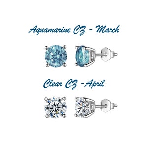 May include: Two pairs of silver stud earrings with clear cubic zirconia stones. The top pair features aquamarine blue stones and the text "Aquamarine CZ - March". The bottom pair features clear stones and the text "Clear CZ - April".