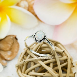 May include: A silver ring with a white opal gemstone set in a delicate, twisted band. The ring is displayed on a woven basket.