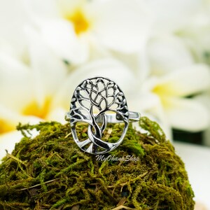 May include: A silver ring with a tree of life design in an oval shape. The ring is displayed on green moss, with white flowers in the background. The shop name "MyOhana Shop" is visible on the moss.