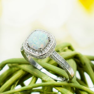 May include: A silver ring with a white opal gemstone surrounded by a halo of smaller clear stones. The ring is set on a green background.