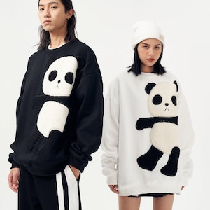 May include: Black and white sweatshirts featuring a panda bear design. One sweatshirt is black with a white panda, the other is white with a black panda. The panda design has a textured, plush appearance.