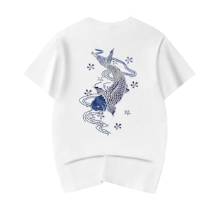 May include: White t-shirt featuring a blue and white koi fish design on the back. The koi fish is surrounded by blue water lines and small white flowers. The shirt has short sleeves and a crew neck.
