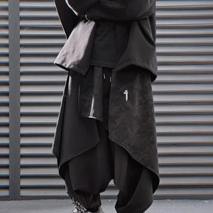 May include: A person wearing a black outfit with a unique design. The outfit includes a hooded top, a draped layer, and loose-fitting pants. The pants have a camouflage pattern on the front and a white graphic. The person is wearing gray and white sneakers.