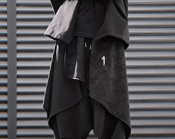 Könnte beinhalten: Eine Person in einem schwarzen Outfit mit einem einzigartigen Design. Das Outfit umfasst einen Kapuzenpullover, eine drapierte Schicht und eine locker sitzende Hose. Die Hose hat ein Camouflagemuster auf der Vorderseite und eine weiße Grafik. Die Person trägt grau-weiße Turnschuhe.