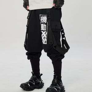 May include: Black cargo pants with white Japanese text and straps. The pants have a unique design with multiple pockets and straps. The outfit includes black sneakers with white accents. The overall style is modern and urban.