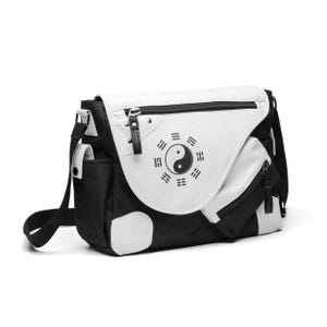 Könnte beinhalten: Eine schwarz-weiße Umhängetasche mit einem Yin-Yang-Symbol, das von acht Trigrammen auf der Frontklappe umgeben ist. Die Tasche verfügt über eine Reißverschlusstasche auf der Klappe, eine Seitentasche, einen verstellbaren schwarzen Schultergurt.