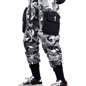 May include: Camouflage print cargo pants in shades of gray, black, and white. The pants feature multiple pockets, black cuffs, and a black pocket with a white graphic. The pants are paired with black and white sneakers.