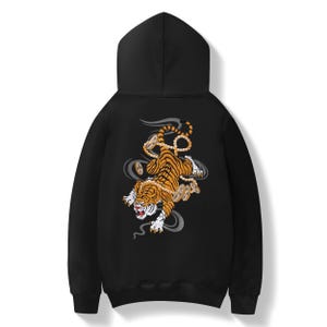 May include: Black hooded sweatshirt with a tiger illustration on the back. The tiger is orange and black, with coins and grey smoke-like swirls. The design is centrally placed on the back of the hoodie.