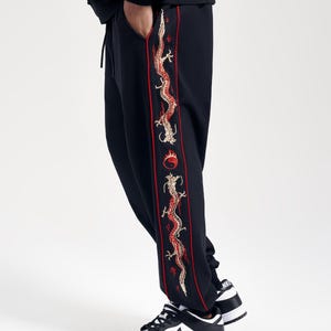 May include: Black sweatpants with a red and gold embroidered dragon design down the side. The pants have a drawstring waist and are paired with black and white sneakers. The dragon design includes a red flame and a yin-yang symbol.