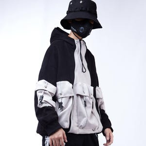 Puede incluir: Chaqueta con capucha negra y blanca con múltiples bolsillos de color gris claro y un sombrero de pescador negro. La chaqueta tiene una cremallera frontal y un diseño único. La persona también lleva una mascarilla negra.