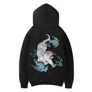 Puede incluir: Sudadera con capucha negra con un diseño de tigre blanco y detalles en azul. El tigre está bordado en la espalda, con la boca abierta. El diseño incluye formas azules en forma de nube. Una sudadera con capucha clásica.