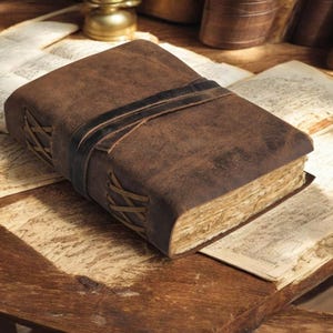 May include: A brown leather journal with a black strap closure, resting on a wooden surface with aged paper. The journal's edges are laced with a lighter-colored thread. The pages appear aged, suggesting a vintage or antique style.