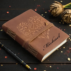 May include: A brown leather journal with a heart-shaped floral design and the name "Michael" embossed on the cover. The journal is secured with a leather strap and sits next to a black pen with gold accents.