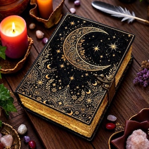 May include: A black hardcover book featuring an intricate gold foil design of a crescent moon, stars, and botanical patterns on the front cover. The book includes a metallic latch closure and has gold-edged pages along the side.