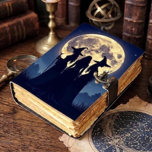 May include: A rectangular journal with a dark blue cover featuring a silhouette of three figures in pointed hats standing before a large, glowing full moon. The book has aged, deckled-edge pages and a metal latch closure on the side.