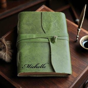Könnte beinhalten: Ein grünes Lederjournal mit Wickelverschluss, personalisiert mit dem Namen "Michelle" in schwarzer Schrift. Eine dekorative Feder und eine kleine Tasse dunkler Flüssigkeit befinden sich in der Nähe, zusammen mit einem dekorativen Stift.