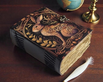 Op de afbeelding: Een decoratief notitieboek met een gedetailleerde illustratie van een konijn op de omslag. De zwarte omslag heeft gouden accenten en een metalen sluiting. De pagina's hebben een verouderde uitstraling, en een witte veer ligt ernaast.