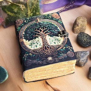 Op de afbeelding: Een decoratief dagboek met een gedetailleerd levensboomontwerp op de omslag. De omslag is donkerblauw met groene accenten en een metalen sluiting. De pagina's hebben een vintage, verouderde uitstraling. Het dagboek is omgeven door kleine stenen.