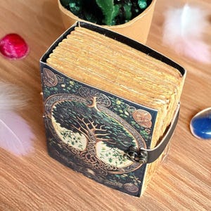 Op de afbeelding: Een decoratief dagboek met een levensboomontwerp op de omslag. De omslag is donkergroen met gouden accenten en een zwarte leren riem. De pagina's zijn verouderd en het dagboek wordt gesloten met een metalen sluiting.