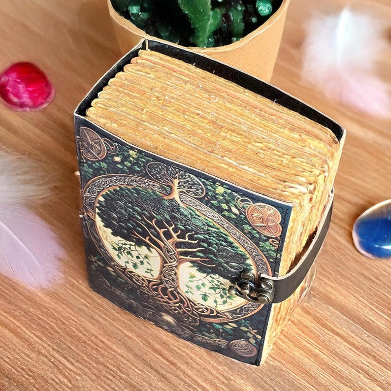 Op de afbeelding: Een decoratief dagboek met een levensboomontwerp op de omslag. De omslag is donkergroen met gouden accenten en een zwarte leren riem. De pagina's zijn verouderd en het dagboek wordt gesloten met een metalen sluiting.