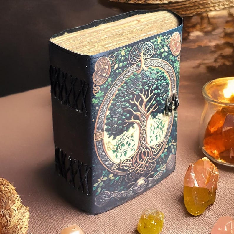 Op de afbeelding: Een donkerblauw leren gebonden dagboek met een levensboomontwerp op de omslag. De omslag heeft een ovaal ontwerp met een boom, Keltische knopen en groen gebladerte. De rug is geregen met zwarte draad en de pagina's zijn verouderd.