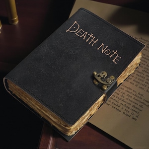 Peut inclure: Un journal en cuir noir avec les mots "DEATH NOTE" en relief sur la couverture. Le journal a des pages jaunies et vieilles et un fermoir en métal. Le livre est ouvert sur une surface avec du texte.