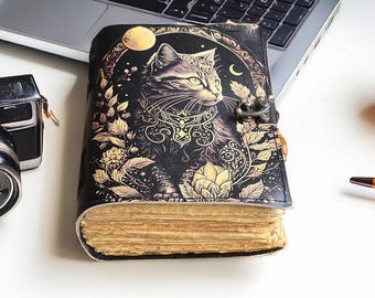 May include: Black leather journal with a cat illustration. The cover features a detailed design of a cat with a moon and floral accents in gold. The journal has a metal clasp and aged-looking pages, suggesting a vintage or fantasy theme.