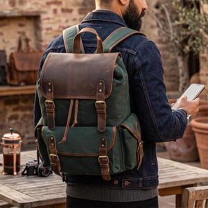 Può includere: Uno zaino in tela verde scuro con patta superiore e finiture delle tasche frontali in pelle marrone. La borsa presenta due cinghie verticali in pelle con fibbie color ottone sul compartimento principale, una chiusura a coulisse e due tasche laterali.