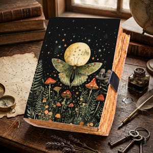 May include: A decorative journal with a black cover featuring a whimsical design of a moth with a moon body, surrounded by stars and mushrooms. The journal has a clasp closure and aged-looking pages, suggesting a vintage or fantasy theme.