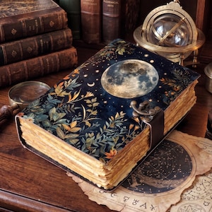 May include: A hardcover book with a dark blue cover featuring a large full moon, golden stars, and green and orange botanical illustrations. The book has a metal latch closure and thick, deckled-edge pages that appear aged and textured.