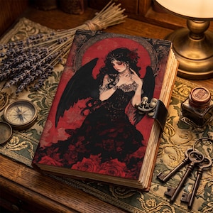 May include: A rectangular journal with a dark red cover featuring an illustration of a figure with black wings and long dark hair surrounded by red roses. The journal has a metal latch closure and aged paper pages visible along the side edge.