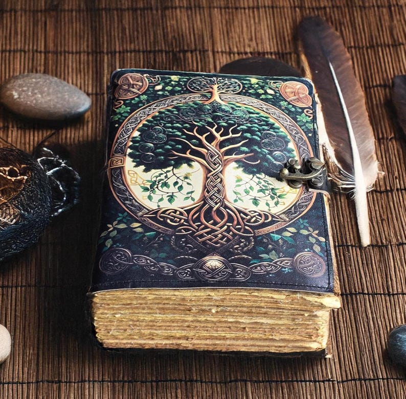 Op de afbeelding: Een donkerblauw leren notitieboek met een levensboomontwerp op de kaft. De kaft heeft een messing sluiting en Keltische knoopwerk. De pagina's hebben een vintage uitstraling en een veer ligt in de buurt.