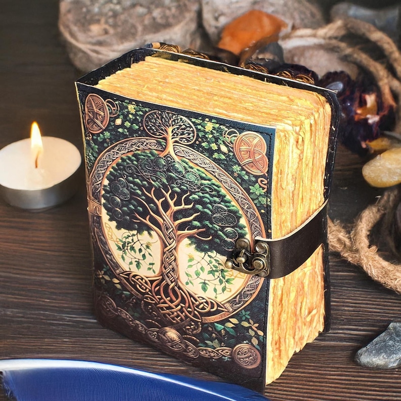 Op de afbeelding: Een leren notitieboek met een levensboomontwerp. De omslag toont een gedetailleerde illustratie van een boom met Keltische knopen. De pagina's hebben een vintage, verouderde uitstraling en een leren band sluit het notitieboek af. Hoogte ongeveer 15 cm.