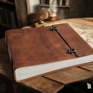 May include: A large, brown leather journal with a clasp closure. The journal is open, revealing the white pages inside. The journal is on a wooden table. The journal is a classic and timeless design.