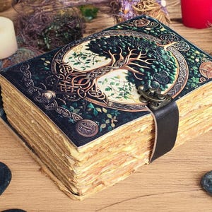Op de afbeelding: Een vintage-stijl dagboek met een levensboomontwerp op de kaft. De kaft is donkerblauw met groene en gouden accenten, en een zwarte leren band sluit het boek. De verouderde pagina's en decoratieve elementen geven het een mystieke uitstraling.