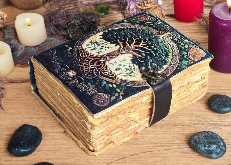Op de afbeelding: Een vintage-stijl dagboek met een levensboomontwerp op de kaft. De kaft is donkerblauw met groene en gouden accenten, en een zwarte leren band sluit het boek. De verouderde pagina's en decoratieve elementen geven het een mystieke uitstraling.
